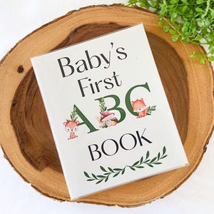 Puede incluir: Un libro blanco "Baby's First ABC Book" con ilustraciones de animales del bosque y detalles de follaje verde, sobre una rodaja de madera. El t&iacute;tulo del libro es en negro, con las letras "ABC" en verde.