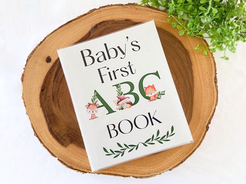 Puede incluir: Un libro blanco "Baby's First ABC Book" con ilustraciones de animales del bosque y detalles de follaje verde, sobre una rodaja de madera. El t&iacute;tulo del libro es en negro, con las letras "ABC" en verde.