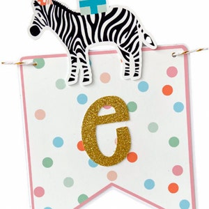 May include: A white banner with a pink border and colorful polka dots. A gold glitter letter "e" is centered on the banner. A zebra with a party hat and gifts is attached to the top of the banner. The banner is strung with gold and white twine.