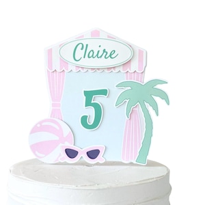 May include: A cake topper featuring a light blue and pink cabana design with the name Claire in green text, a large green number 5, a pink and white beach ball, a pair of pink sunglasses, and a green palm tree silhouette on a white cake.