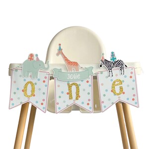 May include: A high chair decorated with a banner for a first birthday. The banner spells "ONE" with glitter letters and features animal illustrations of an elephant, giraffe, and zebra. The banner also has the name "Jolie".