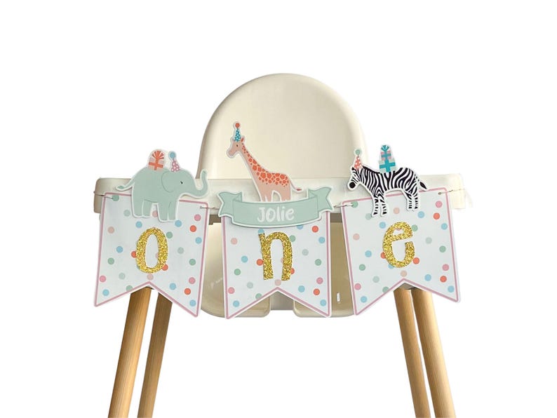 May include: A high chair decorated with a banner for a first birthday. The banner spells "ONE" with glitter letters and features animal illustrations of an elephant, giraffe, and zebra. The banner also has the name "Jolie".