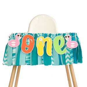 May include: A turquoise high chair skirt with a white and blue chevron pattern. A colorful banner with the word "one" and pink flamingo inflatables hangs from the skirt.
