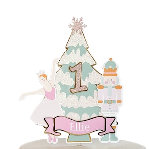 May include: A festive cake topper featuring a ballerina, a nutcracker, and a Christmas tree with a snowflake. The number 1 is displayed on the tree, and a banner reads "Ellie". The design incorporates pink, white, gold, and mint green.