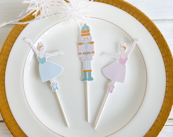 May include: Three decorative cupcake toppers featuring a ballerina and a nutcracker. The ballerina toppers are in light blue and pink, with white arms and legs. The nutcracker is in pink, green, and orange. All are on white sticks, presented on a white plate with a gold rim.