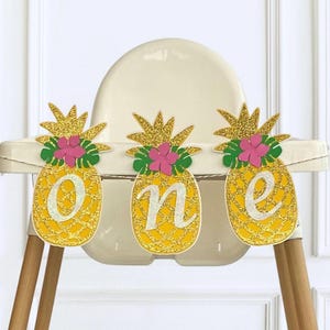 May include: A festive banner with three pineapple cutouts spelling "one." Each yellow pineapple has a gold glitter crown, pink flower, and green leaves. The banner is hanging on a high chair, perfect for a first birthday celebration.