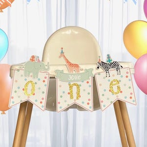 May include: A high chair decorated for a first birthday celebration. The banner reads "One" with animal illustrations and the name "Jolie". Colorful balloons in pink, blue, and yellow surround the chair, creating a festive atmosphere.