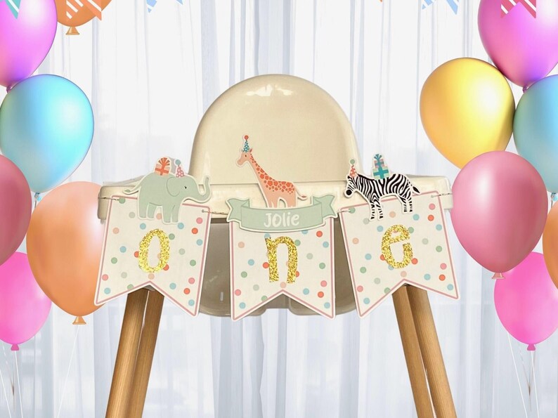 May include: A high chair decorated for a first birthday celebration. The banner reads "One" with animal illustrations and the name "Jolie". Colorful balloons in pink, blue, and yellow surround the chair, creating a festive atmosphere.