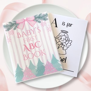 May include: A rectangular book titled Baby's First ABC Book with a pink and white striped cover, featuring a pink bow, green pine branches, snowflakes, and pink and green pine trees. An open page shows the text A is for Angel with an illustration.