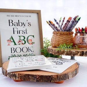 Puede incluir: Un letrero enmarcado con el texto "Baby's First ABC Book" se muestra junto con libros para colorear y crayones. El letrero tiene un marco de madera y presenta ilustraciones de animales del bosque. Los crayones est&aacute;n en una cesta tejida y un tarro de cristal decorativo.