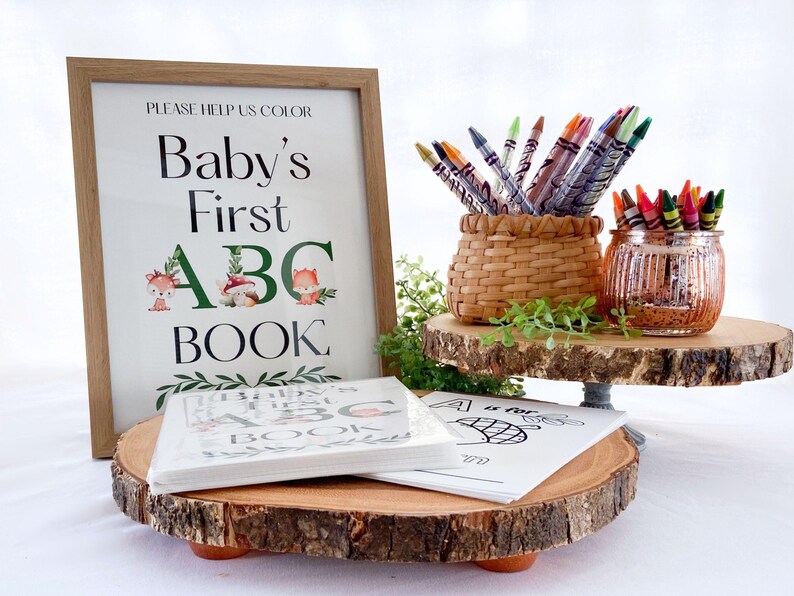 Puede incluir: Un letrero enmarcado con el texto "Baby's First ABC Book" se muestra junto con libros para colorear y crayones. El letrero tiene un marco de madera y presenta ilustraciones de animales del bosque. Los crayones est&aacute;n en una cesta tejida y un tarro de cristal decorativo.