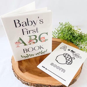 Puede incluir: Un libro blanco "Baby's First ABC Book" con ilustraciones de animales del bosque y una tarjeta flash "A is for acorn" a juego. El libro y la tarjeta se muestran sobre una rodaja de madera con vegetaci&oacute;n.