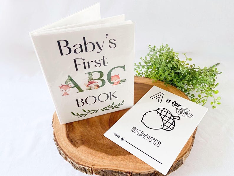 Puede incluir: Un libro blanco "Baby's First ABC Book" con ilustraciones de animales del bosque y una tarjeta flash "A is for acorn" a juego. El libro y la tarjeta se muestran sobre una rodaja de madera con vegetaci&oacute;n.