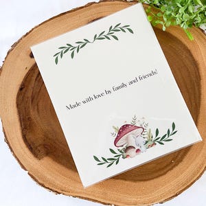Puede incluir: Una tarjeta blanca con el texto "Made with love by family and friends!" se muestra sobre una superficie de madera. La tarjeta presenta una ilustraci&oacute;n de acuarela de setas y un peque&ntilde;o animal. Acentos de hojas verdes enmarcan la parte superior e inferior.