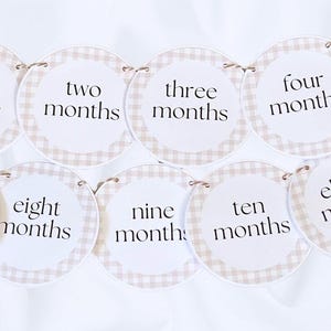 May include: A circular banner featuring beige and white gingham borders with black text indicating months from one to eleven. Each round card is connected by thin string, displaying phrases like one month, two months, three months, and four months.