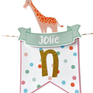 May include: A festive banner with a giraffe wearing a party hat, the name "Jolie" on a banner, and a gold glitter letter "n". The banner is decorated with colorful polka dots and a pink border, perfect for a birthday celebration.
