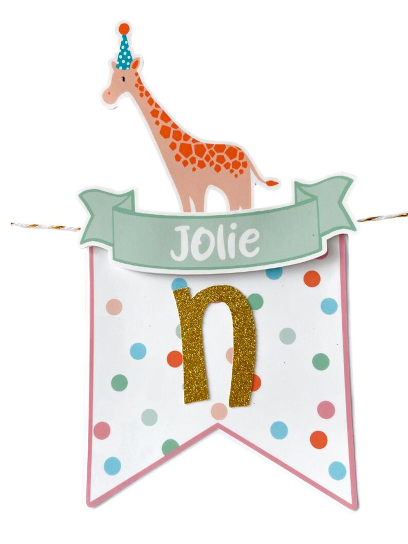 May include: A festive banner with a giraffe wearing a party hat, the name "Jolie" on a banner, and a gold glitter letter "n". The banner is decorated with colorful polka dots and a pink border, perfect for a birthday celebration.