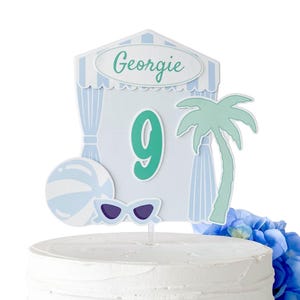 May include: A cake topper featuring a beach cabana design with the name Georgie and the number 9 in teal. The decoration includes a light blue and white striped beach ball, a pair of light blue sunglasses, and a teal palm tree silhouette.