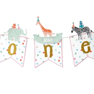 May include: A festive banner for a first birthday celebration. The banner features three pennants with the word "ONE" spelled out in gold glitter letters. Each pennant has a different animal on top: an elephant, a giraffe, and a zebra.