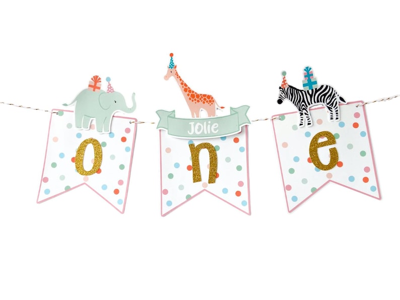 May include: A festive banner for a first birthday celebration. The banner features three pennants with the word "ONE" spelled out in gold glitter letters. Each pennant has a different animal on top: an elephant, a giraffe, and a zebra.
