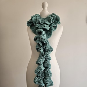 May include: A teal blue crocheted scarf with a ruffled design. The scarf is displayed on a white mannequin.