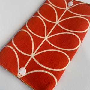May include: A rectangular fabric wallet featuring a vibrant orange background with a repeating white line-drawn leaf pattern. The wallet is secured with two light pink plastic snap buttons positioned at the top and bottom edges of the front flap.