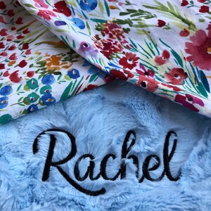 May include: A light blue plush blanket with the name "Rachel" embroidered in black cursive lettering. The blanket is folded over a white fabric with a floral pattern.