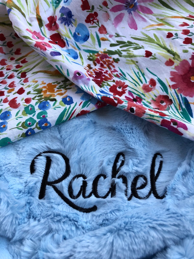 May include: A light blue plush blanket with the name "Rachel" embroidered in black cursive lettering. The blanket is folded over a white fabric with a floral pattern.