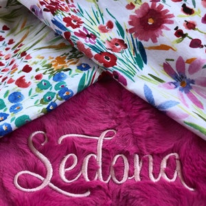 May include: A pink plush blanket with the name "Sedona" embroidered in a cursive font. The blanket is folded over a white fabric with a floral pattern.
