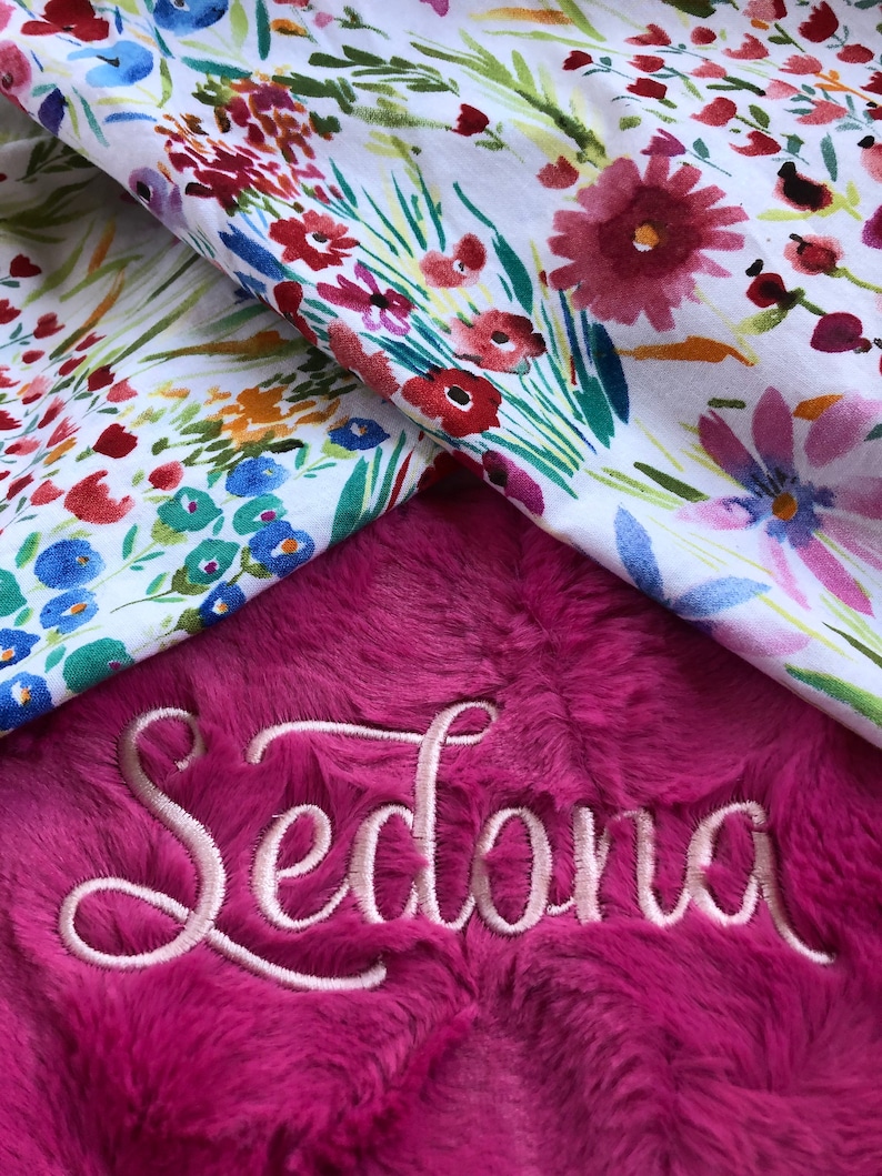 May include: A pink plush blanket with the name "Sedona" embroidered in a cursive font. The blanket is folded over a white fabric with a floral pattern.