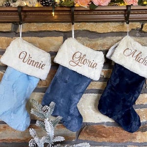May include: Three plush Christmas stockings in light blue, medium blue, and dark navy blue, each featuring a white faux fur cuff. The cuffs are embroidered with the names Vinnie, Gloria, and Michael in a brown script font, hanging from hooks.