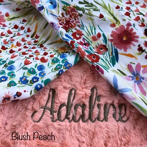May include: A blush peach faux fur baby blanket with the name "Adaline" embroidered in brown thread. The blanket is folded over a white fabric with a floral pattern.