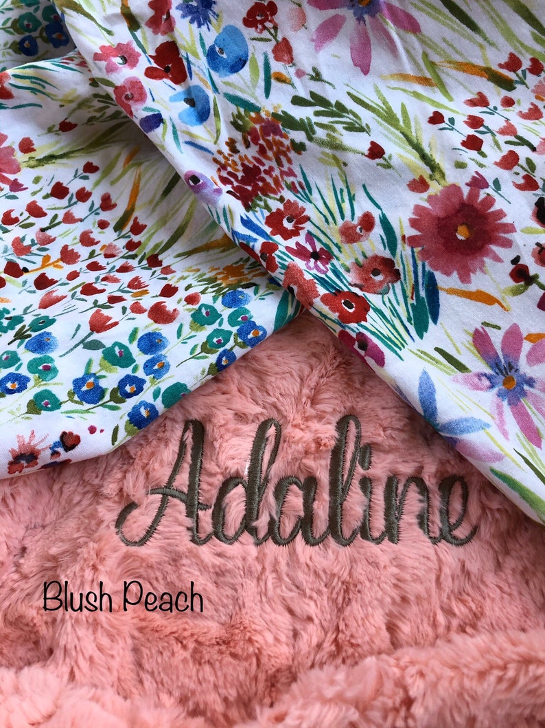 May include: A blush peach faux fur baby blanket with the name "Adaline" embroidered in brown thread. The blanket is folded over a white fabric with a floral pattern.