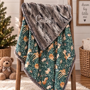 May include: A soft blanket featuring a dark green background with a repeating pattern of woodland animals including deer, foxes, and rabbits among foliage. The top corner is folded over to reveal a dark gray faux fur lining with the name Carter embroidered.