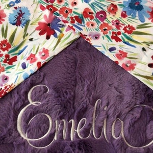 May include: A purple plush blanket with the name "Emelia" embroidered in white thread. The blanket is decorated with a floral pattern.