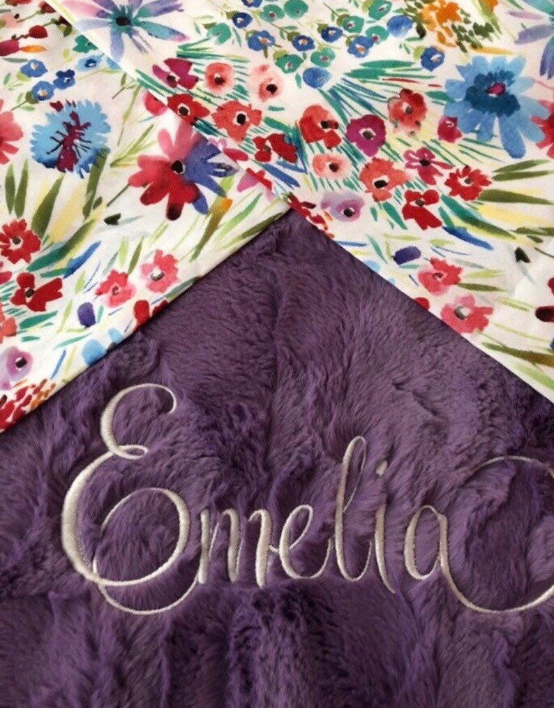 May include: A purple plush blanket with the name "Emelia" embroidered in white thread. The blanket is decorated with a floral pattern.