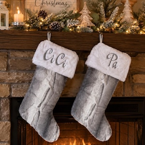 May include: Two gray and white faux fur Christmas stockings hanging from a fireplace mantel. Each stocking features a plush white cuff with embroidered lettering. The left stocking is embroidered with CiCi and the right stocking with Pa.
