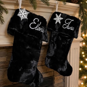 May include: Two black faux fur Christmas stockings featuring a white embroidered name on the cuff. The left stocking is personalized with the name Elana and the right with Juna. Each stocking is accented with a white snowflake ornament.