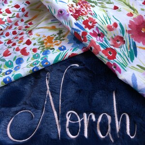 May include: A navy blue plush blanket with the name "Norah" embroidered in white. The blanket is folded over a white fabric with a floral pattern.