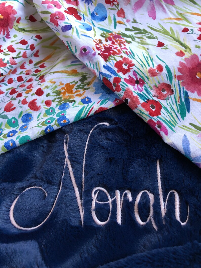 May include: A navy blue plush blanket with the name "Norah" embroidered in white. The blanket is folded over a white fabric with a floral pattern.