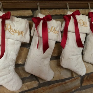 May include: Four white faux fur Christmas stockings hanging in a row. Each stocking features a folded cuff embroidered with a name in gold thread and is decorated with a dark red satin ribbon bow tied at the top corner of the cuff.