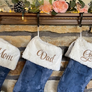 May include: Three Christmas stockings with deep blue plush bodies and white faux fur cuffs. Each cuff features embroidered text in a metallic copper script font reading Mom, Dad, and Gloria, respectively, hanging from black metal hooks.