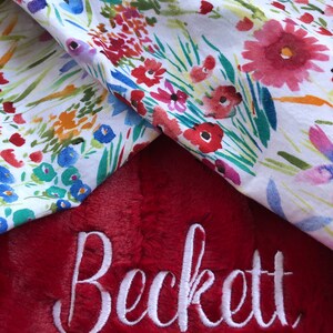 May include: A red plush blanket with the name "Beckett" embroidered in white. The blanket is folded over a white fabric with a floral pattern.