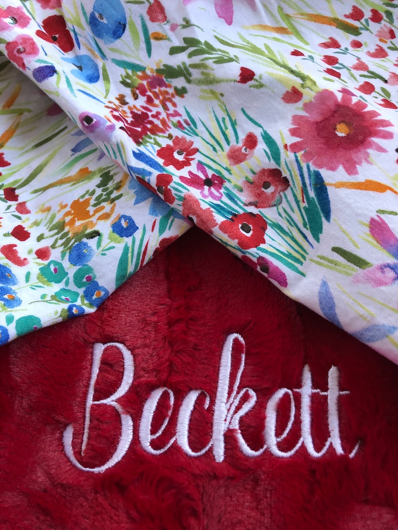 May include: A red plush blanket with the name "Beckett" embroidered in white. The blanket is folded over a white fabric with a floral pattern.
