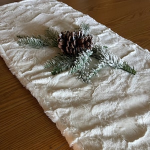 May include: White faux fur table runner with a pine sprig and pine cone on top. The runner has a textured, fluffy surface.