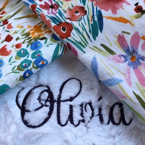 May include: A white plush blanket with the name "Olivia" embroidered in black cursive lettering. The blanket is folded over a floral fabric with a white background and colorful flowers.