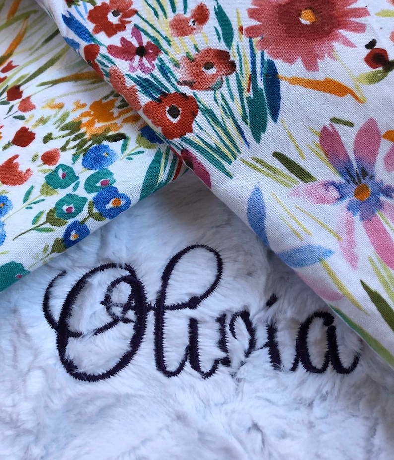 May include: A white plush blanket with the name "Olivia" embroidered in black cursive lettering. The blanket is folded over a floral fabric with a white background and colorful flowers.