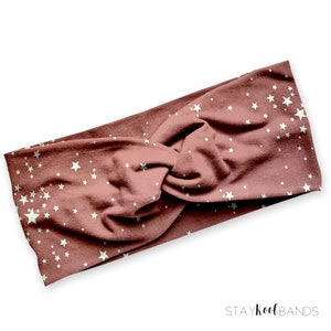 May include: A dusty rose-colored headband with a white star pattern. The fabric is twisted at the center. The text "STAYKOOLBANDS" is printed in the bottom right corner. This accessory is suitable for various hairstyles.
