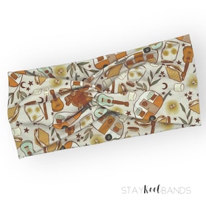 May include: A white headband with a pattern of camping-themed illustrations, including a camper van, campfire, guitar, and s'mores. The text "STAY Kool BANDS" is printed on the bottom of the image.