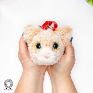 May include: A plush cat head ornament with a red ribbon, blue eyes, and a white nose. The ornament is surrounded by red Christmas ornaments.